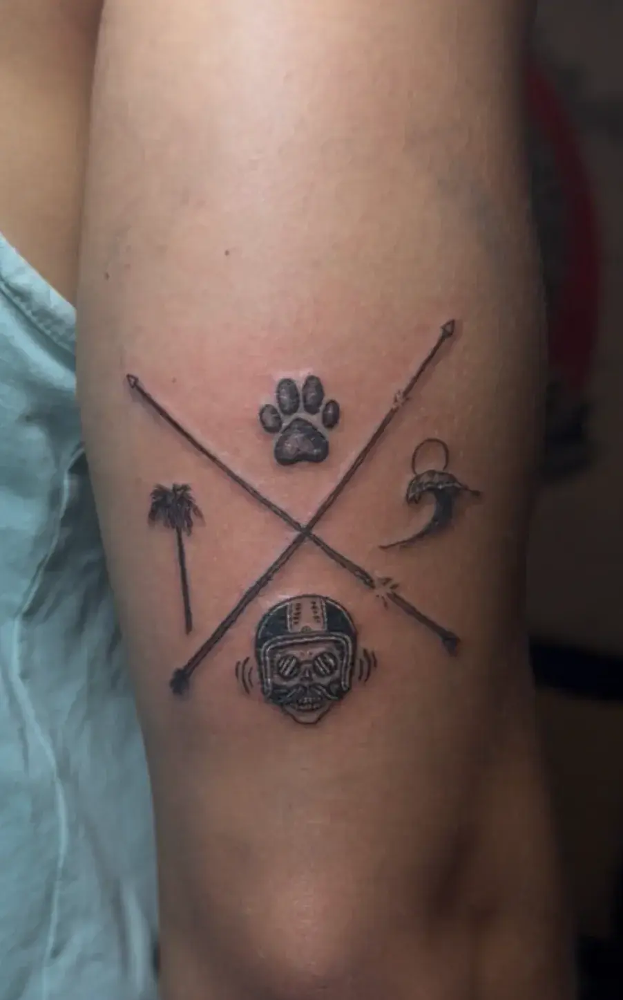 Tatuaje pequeño de flechas cruzadas con huella, palmera, ola y calavera con casco en el brazo
